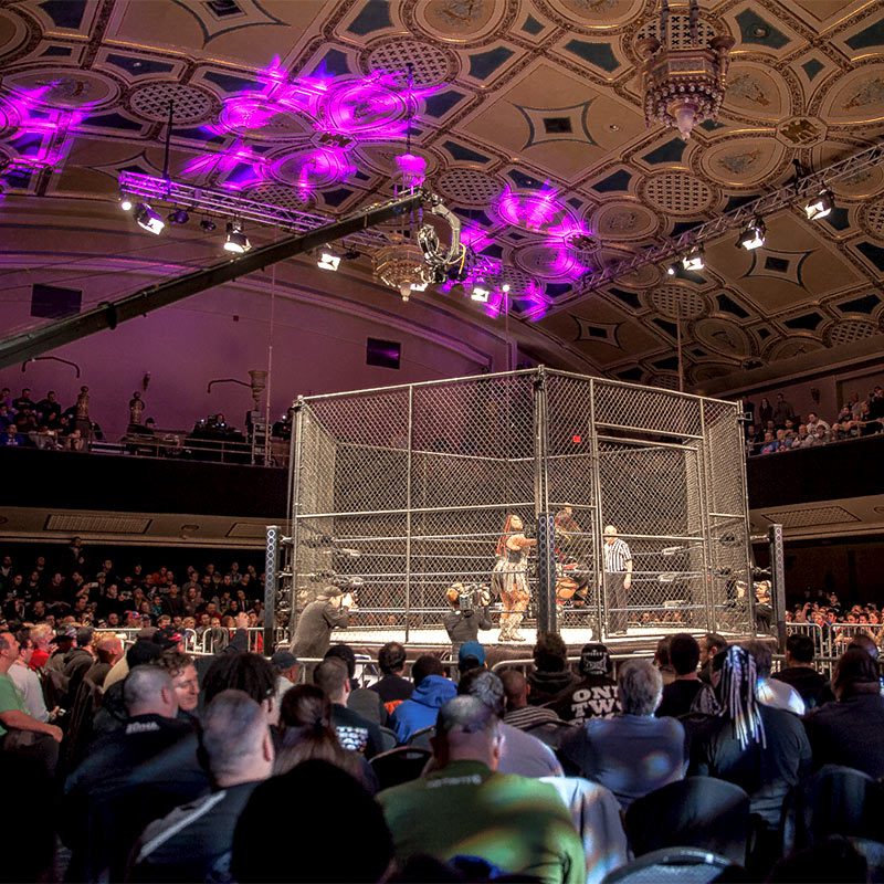 A large crowd gathers in an arena, intently watching an exciting wrestling match unfold in the center ring.